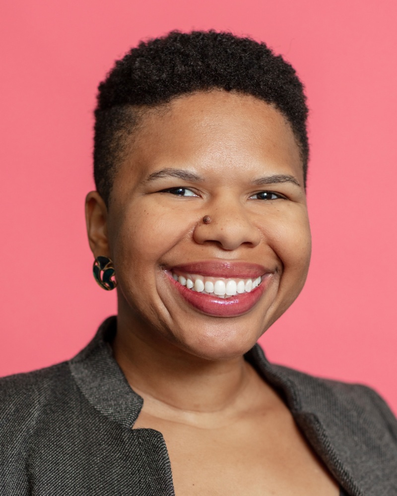 A person with short hair, wearing a gray jacket and earrings, smiles in front of a pink background.